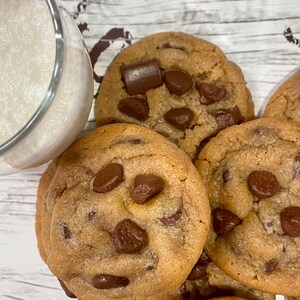 May include: A close-up of several freshly baked chocolate chip cookies with large chocolate chunks. A glass of milk is partially visible in the upper left corner. The cookies are golden brown and appear soft.