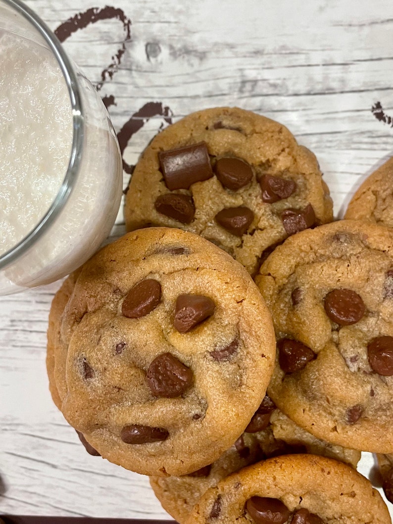 May include: A close-up of several freshly baked chocolate chip cookies with large chocolate chunks. A glass of milk is partially visible in the upper left corner. The cookies are golden brown and appear soft.
