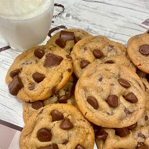 May include: A stack of freshly baked chocolate chip cookies with large chocolate chunks. A glass of milk sits next to the cookies. The cookies are golden brown and appear soft and chewy.