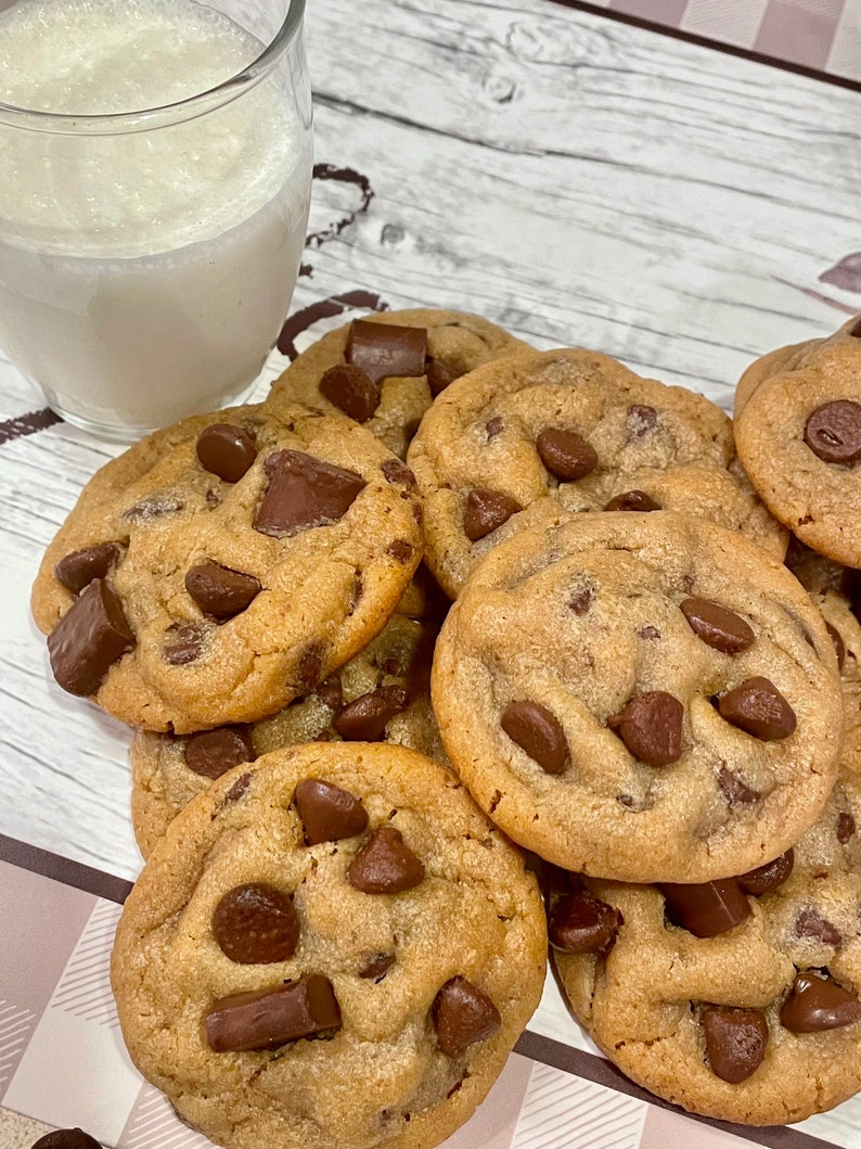 May include: A stack of freshly baked chocolate chip cookies with large chocolate chunks. A glass of milk sits next to the cookies. The cookies are golden brown and appear soft and chewy.