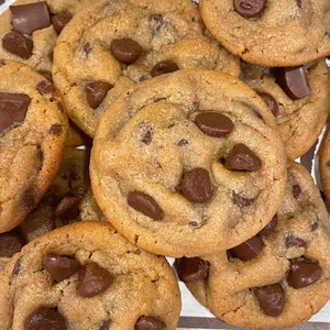 May include: A close-up shot of several freshly baked chocolate chip cookies. The cookies are golden brown with melted chocolate chips and chunks throughout. The cookies are stacked on top of each other.