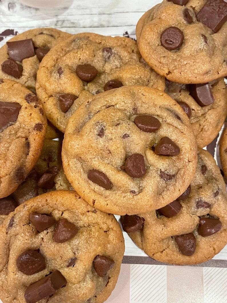 May include: A close-up shot of several freshly baked chocolate chip cookies. The cookies are golden brown with melted chocolate chips and chunks throughout. The cookies are stacked on top of each other.