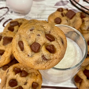 May include: Close-up of several freshly baked chocolate chip cookies with large chocolate chunks. One cookie rests on the rim of a small glass of milk. The cookies are golden brown and sit on a pink and white checkered surface.