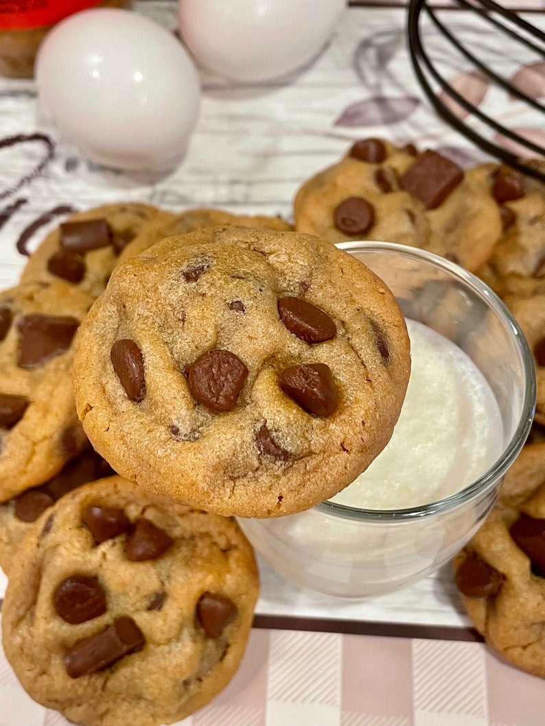May include: Close-up of several freshly baked chocolate chip cookies with large chocolate chunks. One cookie rests on the rim of a small glass of milk. The cookies are golden brown and sit on a pink and white checkered surface.
