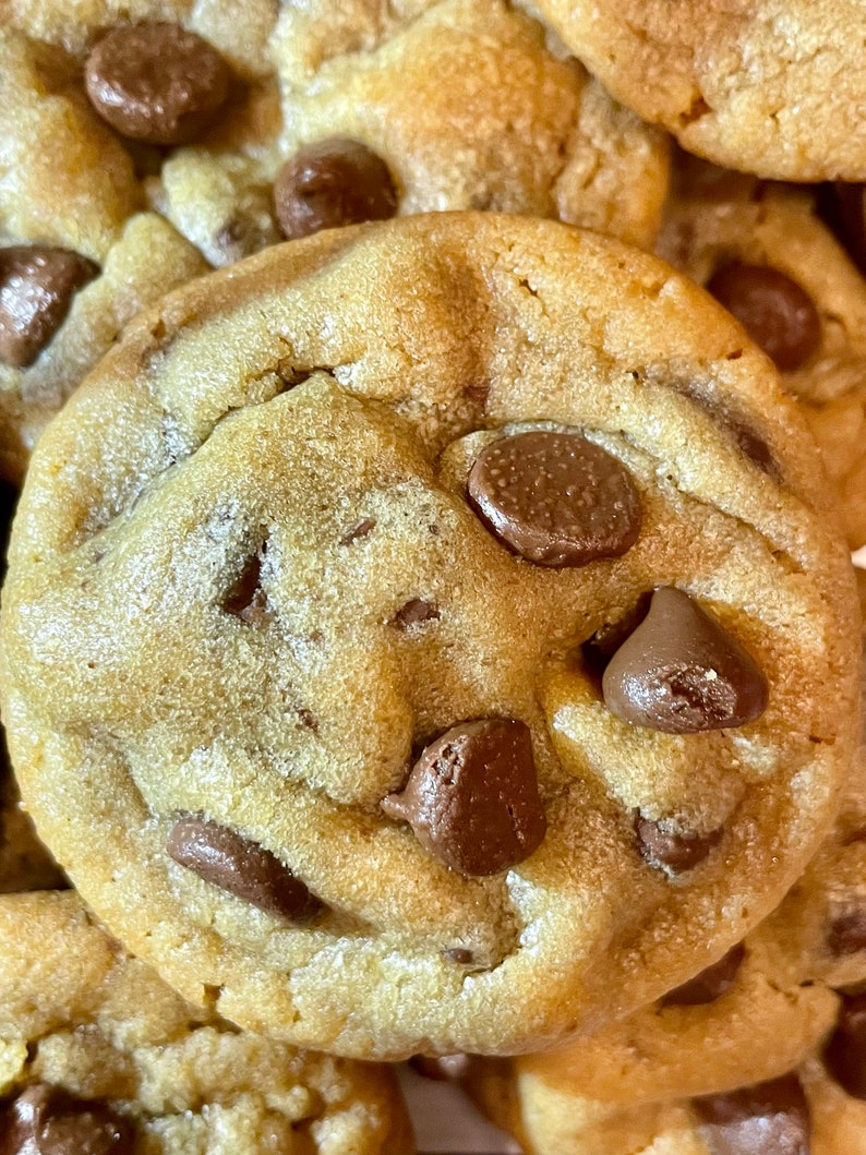 May include: Close-up of a golden-brown chocolate chip cookie with several chocolate chips. The cookie has a slightly uneven texture and is surrounded by other cookies.