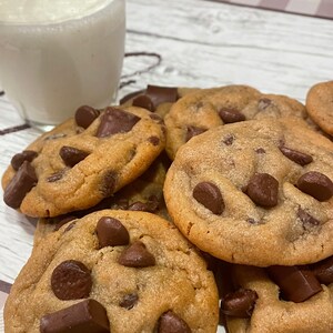 May include: A close-up of several freshly baked chocolate chip cookies with large chocolate chunks. A glass of milk is in the background. The cookies are golden brown and appear soft and chewy.