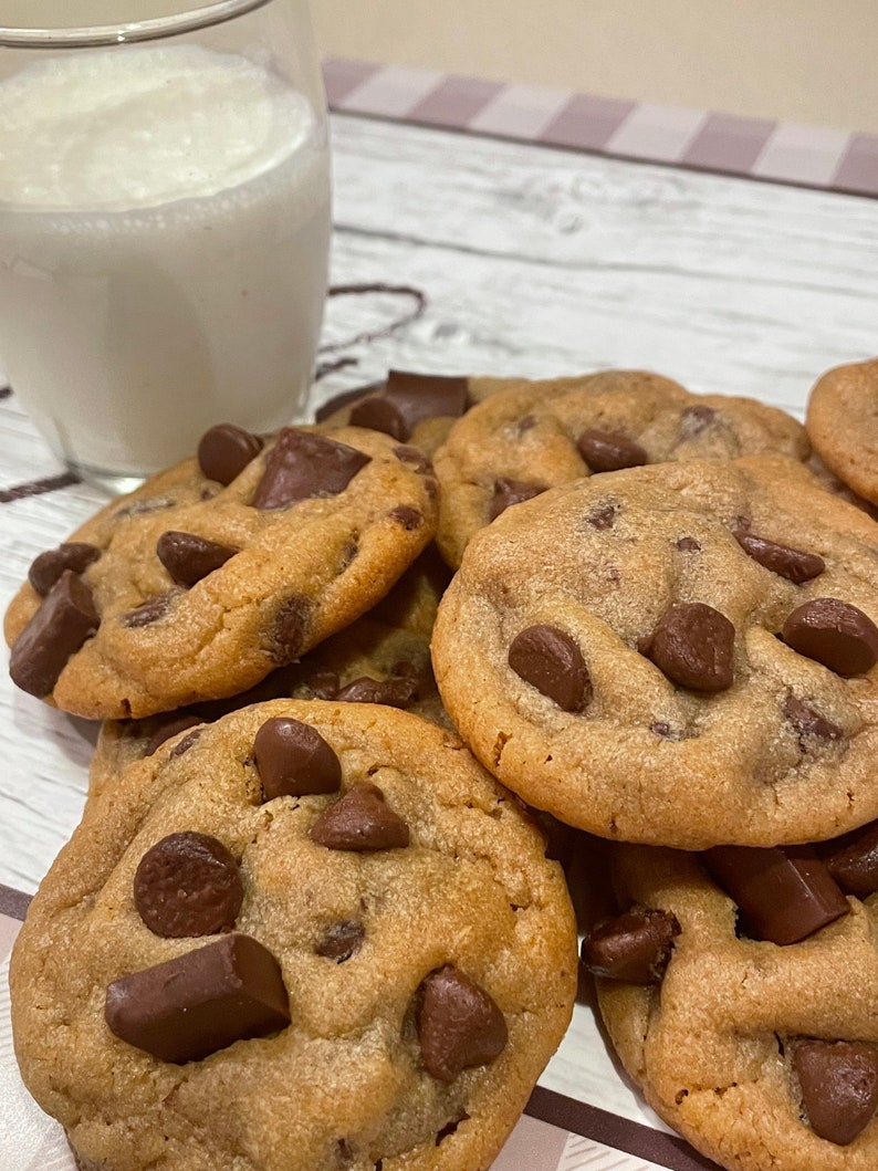 May include: A close-up of several freshly baked chocolate chip cookies with large chocolate chunks. A glass of milk is in the background. The cookies are golden brown and appear soft and chewy.
