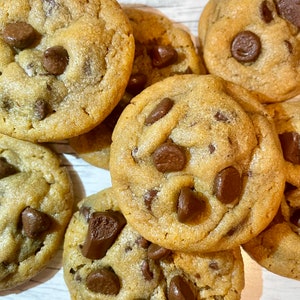 May include: A close-up shot of several freshly baked chocolate chip cookies. The cookies are golden brown with visible chocolate chips scattered throughout. The cookies are arranged on a light-colored surface.