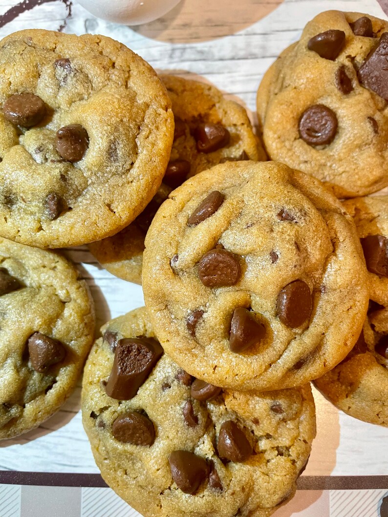 May include: A close-up shot of several freshly baked chocolate chip cookies. The cookies are golden brown with visible chocolate chips scattered throughout. The cookies are arranged on a light-colored surface.