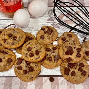 May include: A collection of freshly baked chocolate chip cookies with large chocolate chunks. Two white eggs and a jar of peanut butter are in the background, along with a black whisk. The cookies are arranged on a patterned surface.