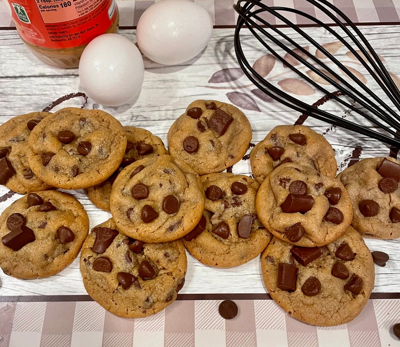 May include: A collection of freshly baked chocolate chip cookies with large chocolate chunks. Two white eggs and a jar of peanut butter are in the background, along with a black whisk. The cookies are arranged on a patterned surface.