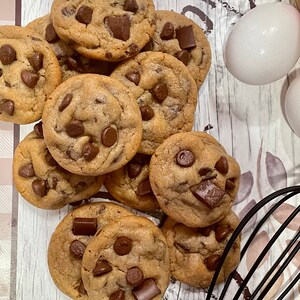 May include: A pile of freshly baked chocolate chip cookies with large chocolate chunks. Two white eggs and a black whisk are visible in the background, suggesting a baking theme. The cookies are golden brown and appear soft.