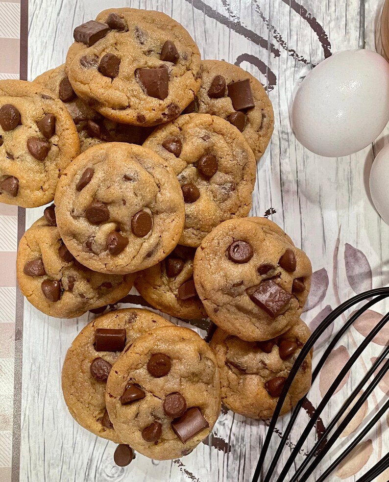 May include: A pile of freshly baked chocolate chip cookies with large chocolate chunks. Two white eggs and a black whisk are visible in the background, suggesting a baking theme. The cookies are golden brown and appear soft.