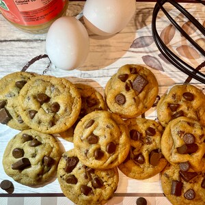 May include: A collection of freshly baked chocolate chip cookies with large chocolate chunks. Two white eggs, a jar of peanut butter, and a black whisk are also visible, suggesting a baking theme.