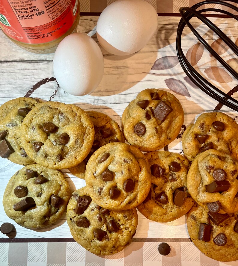 May include: A collection of freshly baked chocolate chip cookies with large chocolate chunks. Two white eggs, a jar of peanut butter, and a black whisk are also visible, suggesting a baking theme.