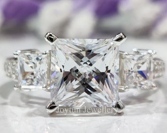 Puede incluir: Un anillo de plata con una piedra central grande de forma cuadrada y dos piedras cuadradas más pequeñas a cada lado. El anillo está colocado sobre un fondo blanco.
