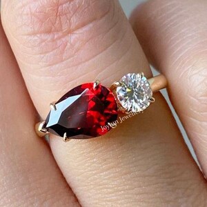 Könnte beinhalten: Ein goldener Ring mit einem birnenförmigen roten Edelstein und einem runden weißen Edelstein. Der Ring wird an einem Finger getragen.