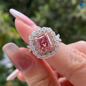 Può includere: Un anello di fidanzamento con diamante rosa con un doppio alone. L'anello è in oro bianco e presenta un grande diamante rosa di forma quadrata al centro. L'alone è composto da diamanti più piccoli e la fascia è anche incastonata con diamanti.