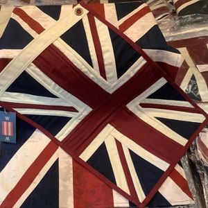May include: A close-up of a red, white and blue Union Jack flag made of fabric. The flag is folded in half and has a white border around the edges. The flag is on a table with other flags in the background.