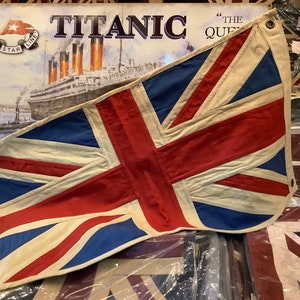 May include: A large, vintage Union Jack flag made of canvas with a white background, red crosses, and blue accents. The flag is hanging from a wall and is slightly faded. The flag is labeled "White Star Line" and "Titanic" in the background.