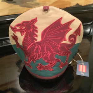 May include: A tea cosy with a beige top and a green bottom, featuring an embroidered burgundy Welsh dragon. The cozy has a tag with the letters "W" and vertical stripes. The cozy is designed to keep tea warm.