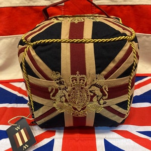 May include: A red, white and blue doorstop with a gold embroidered royal crest and a gold rope trim. The doorstop is shaped like a cube and has a handle on the top.