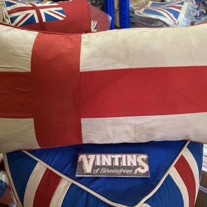 May include: A rectangular throw pillow featuring a red cross on a cream background, resembling the St. George's Cross flag. The pillow has red piping along the edges and a metal grommet on the bottom left corner. A rectangular sign reads VINTINS of Birmingham.