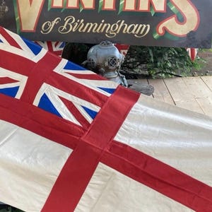 May include: A white flag featuring a red cross and a Union Jack in the upper left corner. The flag is made of fabric and includes a metal grommet at the top corner for hanging. The design is clean and features bold red, white, and blue colours.