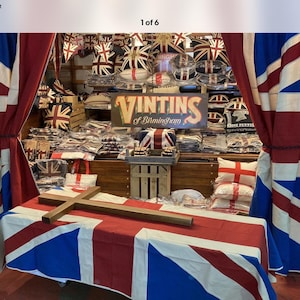 May include: A wooden cross rests on a table covered with a Union Jack flag tablecloth. In the background, a sign reads VINTINS of Birmingham, surrounded by various cushions and textiles featuring the Union Jack and St. George's Cross patterns.