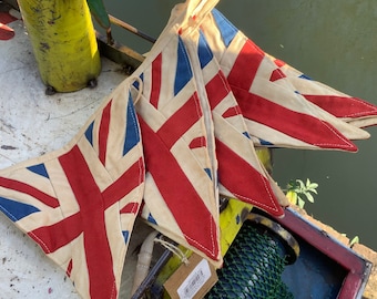 Könnte beinhalten: Eine Girlande aus Stofffahnen mit dem Union-Jack-Design. Die Fahnen sind aus cremefarbenem Stoff mit roten, weißen und blauen Akzenten. Die Girlande hängt an einer Holzoberfläche.
