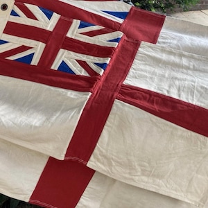 May include: A large fabric flag featuring the Union Jack design in the upper corner with red, white, and blue colours. The flag has a prominent red cross centred on a cream-coloured background, constructed from stitched panels with a metal grommet.