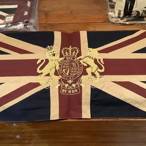 May include: A Union Jack flag-themed decorative item. The flag features a navy blue background with red and white stripes, and a central emblem with gold detailing. The item has a beige border and grommets for hanging. Other similar items are visible in the background.