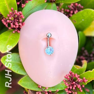 May include: A silver belly button ring with a clear, sparkly gem. The ring is displayed on a pink, flesh-toned surface.