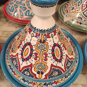 May include: A white ceramic tagine with a blue rim and intricate colorful floral designs. The tagine has a round base and a conical lid with a small opening at the top.