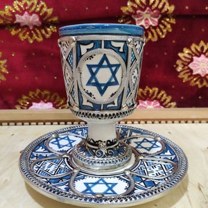 May include: A blue and white ceramic Kiddush cup with a silver rim and a Star of David design. The cup is sitting on a matching plate with a silver rim.