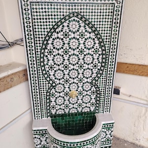 Op de afbeelding: Een decoratieve wandfontein met een mozaïektegelontwerp. De fontein heeft een wit en groen geometrisch patroon, een gebogen bekken en een goudkleurige kraan. Het algehele ontwerp is sierlijk en gedetailleerd.