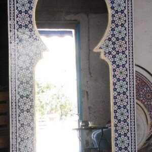 Può includere: Grande specchio rettangolare con una parte superiore ad arco, incorniciato con un motivo a mosaico blu, bianco e oro. Lo specchio riflette una scena esterna luminosa. Il design della cornice è intricato e decorativo.