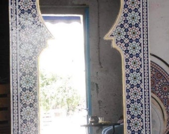 May include: Large rectangular mirror with an arched top, framed with a mosaic tile pattern in blue, white, and gold. The mirror reflects a bright outdoor scene. The frame's design is intricate and decorative.