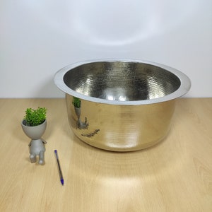 May include: A large, silver, hammered metal bowl with a wide, rounded rim. The bowl is sitting on a light brown wooden surface.