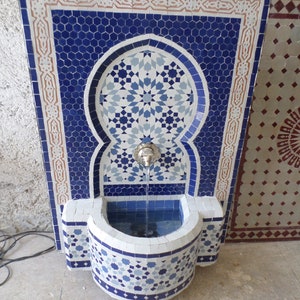 Op de afbeelding: Mozaïektegel fontein met een blauw en wit geometrisch ontwerp. De fontein heeft een centrale boog met een sterrenpatroon, een klein bekken en een zilveren kraan. De tegels zijn in tinten blauw, wit en bruin.
