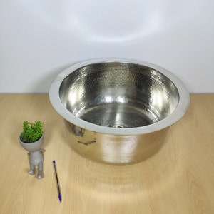May include: A large, round, hammered stainless steel sink with a polished finish. The sink is empty and sits on a light wood surface.