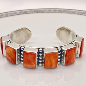 May include: Silver cuff bracelet featuring rectangular orange-red stones. The bracelet has a polished silver finish with a row of small silver beads between each stone. The stones are evenly spaced around the bracelet.