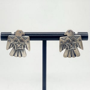 May include: A pair of silver-toned stud earrings. Each earring is shaped like a stylized bird with a star in the center. The earrings are displayed on a black stand against a white background. The bird's wings are spread.