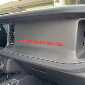 May include: Interior dashboard of a vehicle with the red word "BRONCO" across the center. The dashboard is gray with black trim and the word "AIRBAG" is visible above the red letters.