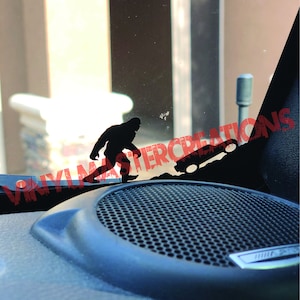 May include: A black silhouette of Bigfoot walking up a hill with a small truck behind him. The image is a sticker for a car window.