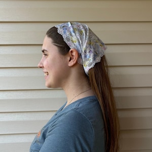 May include: A floral patterned headscarf with a white lace trim. The scarf features a delicate design with pastel colors, including blue, purple, and green. The head covering is tied at the back and worn on the head.