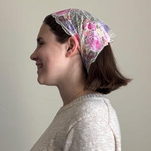 May include: A headscarf with a floral design and a lace overlay. The scarf displays pink, purple, and yellow roses. The head covering is tied at the back and worn on the head.