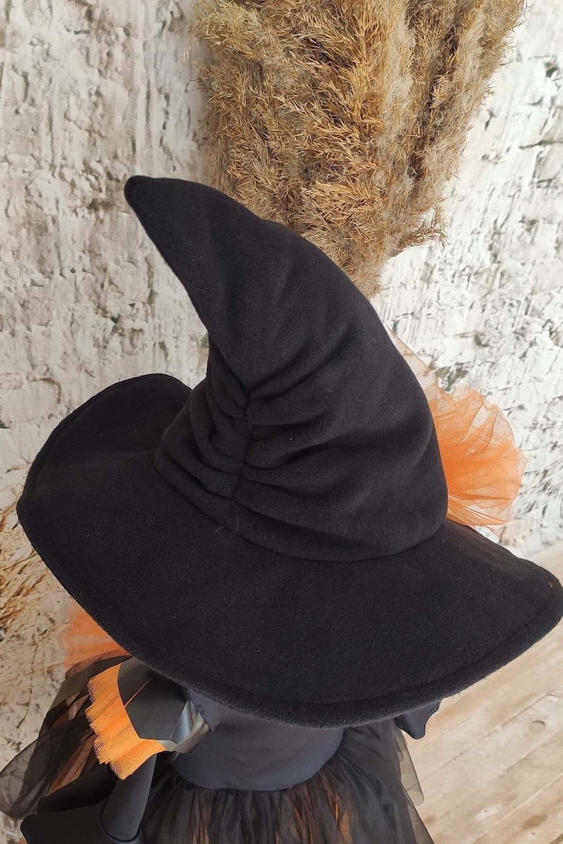 May include: Black witch hat with a tall, pointed crown and a wide brim. The hat is made of soft, plush fabric and has a gathered detail on the crown.