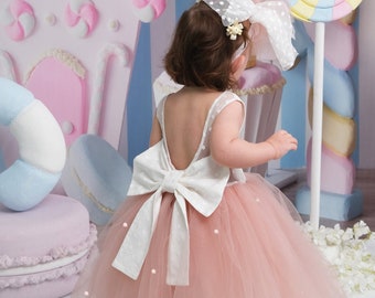 May include: A young girl wearing a pink tulle dress with a white bow and pearl accents. The dress has a low back and is worn with a white tulle headband with a flower accent. The girl is standing in front of a backdrop with a candy-themed design.