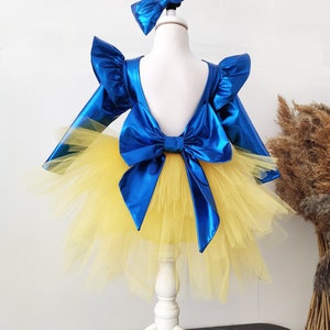 Puede incluir: Un vestido de tul azul y amarillo con un gran lazo azul en la espalda. El vestido tiene mangas largas y un escote con volantes. El vestido est&aacute; en un maniqu&iacute; blanco.